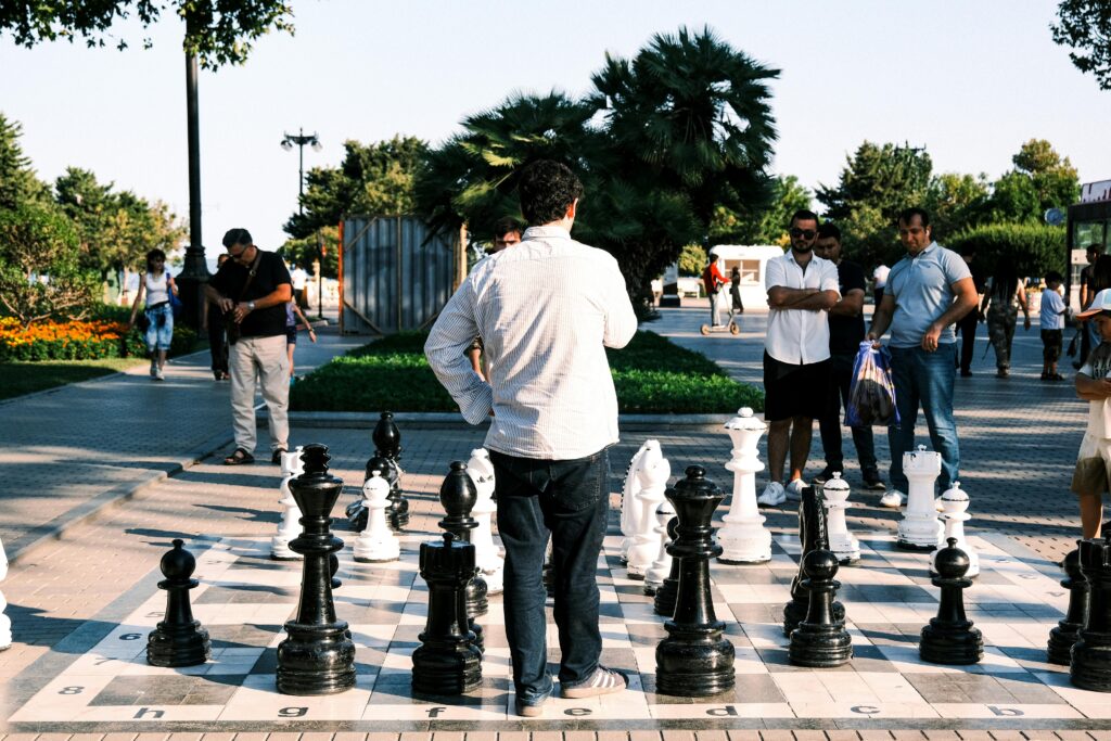 Group of adults playing outdoor chess in a sunny park, surrounded by greenery and spectators.